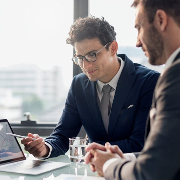 Two businessmen in suits discussing data on a laptop in a modern office setting.