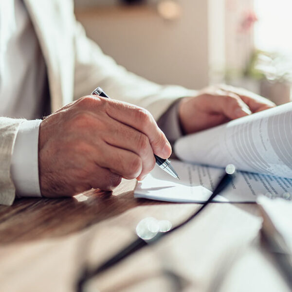 Close-up of a man signing a document at a desk with papers and glasses in view.