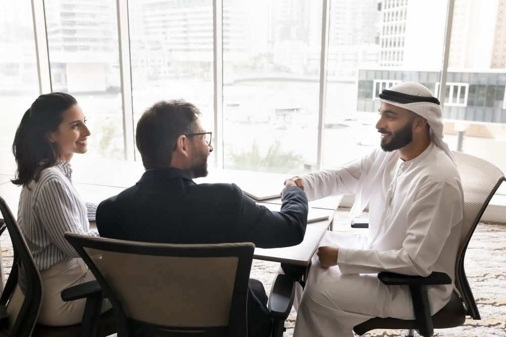 Arab man in kandura shaking hands with businessman at meeting, woman smiling beside them.