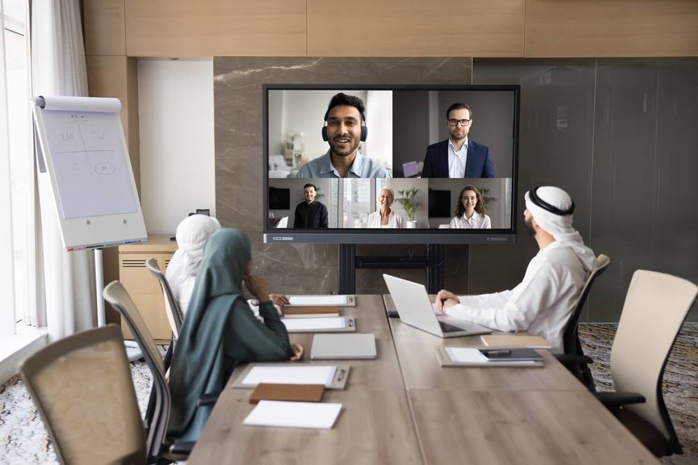 Team in traditional attire in a meeting room joining a video conference on a large screen.